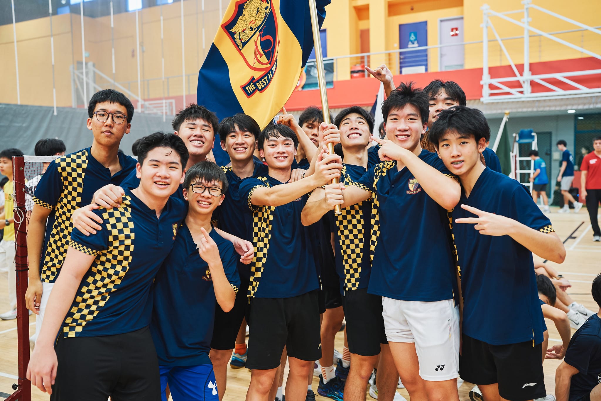 A group of male students in navy and gold checkered uniforms poses energetically for a team photo in a sports hall, making celebratory gestures.